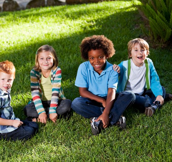 Four kids are sitting on the grass, looking at the camera and smiling.