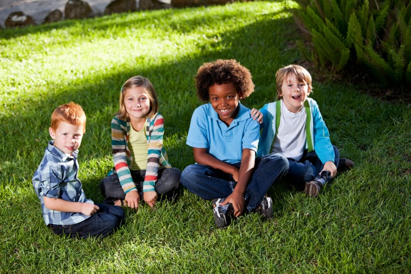 Four kids are sitting on the grass, looking at the camera and smiling.